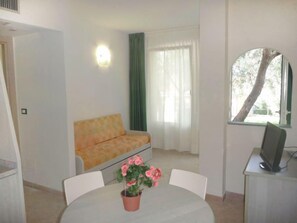 A small, well-lit room with a sofa, a dining table with chairs, a potted plant, and a TV on a stand.