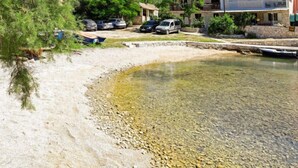 Zona costera con aguas poco profundas y transparentes, costa rocosa, coches aparcados y edificios al fondo.