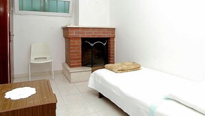 Una habitación pequeña y sencilla con una cama individual, una silla blanca, una mesita y una chimenea de ladrillo.