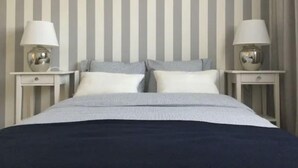 Un lit bien fait, recouvert d’une couvre-lit à rayures, deux tables de chevet avec des lampes et une étagère au-dessus du lit où sont exposées des photos encadrées.