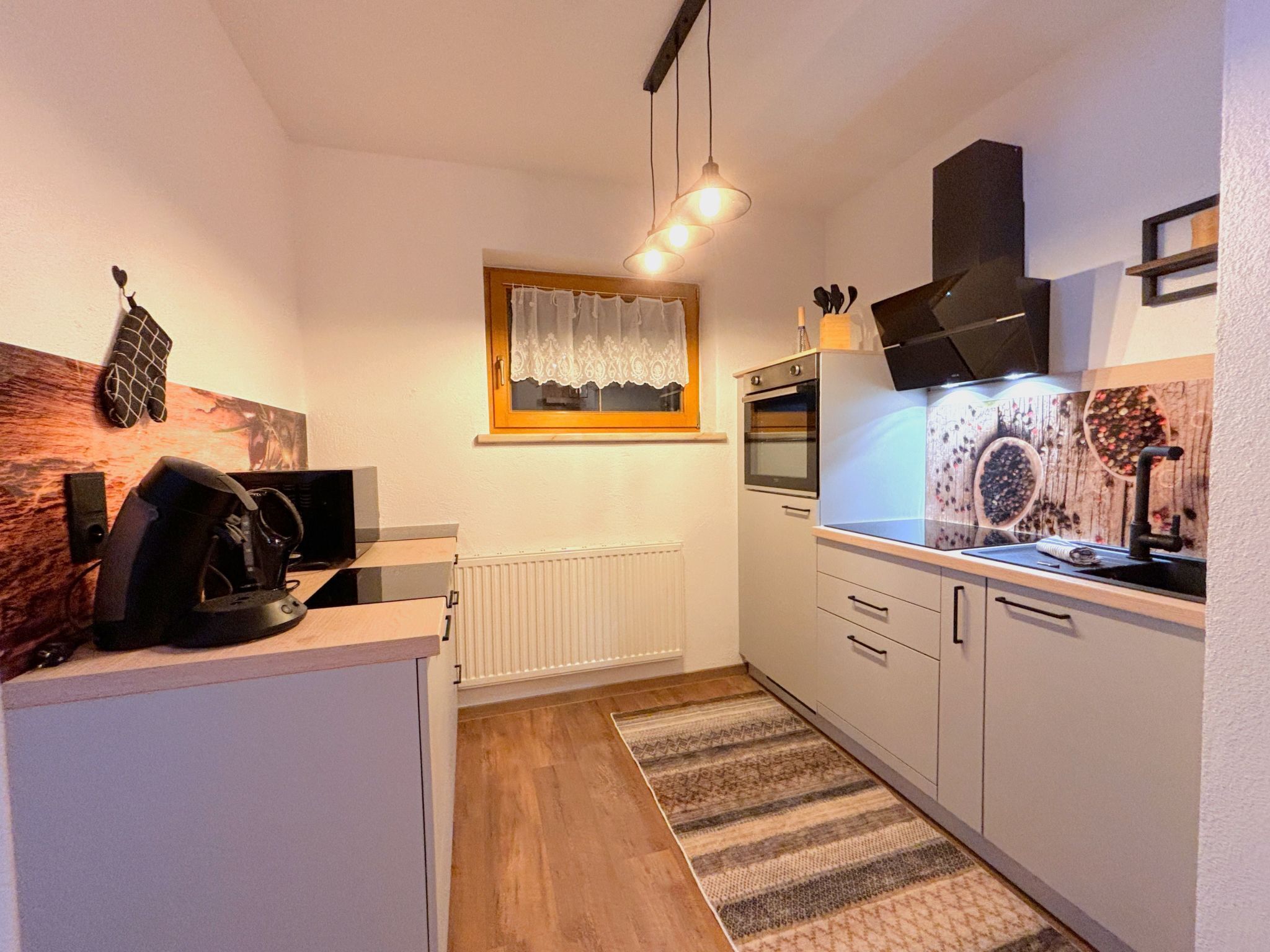 A compact kitchen with white cabinets, a built-in oven, a microwave, and a kettle on the counter. There is a window with a lace curtain, a wooden backsplash, and a patterned rug on the floor.