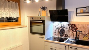 Une cuisine compacte équipée d’un four encastré, d’un micro-ondes et d’un évier. Trois lampes suspendues sont fixées au-dessus du plan de travail, et une fenêtre est agrémentée d’un rideau décoratif.