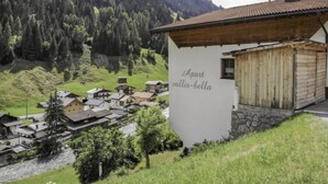 Un village de montagne aux maisons traditionnelles en bois, un bâtiment blanc portant l’inscription « Apart vallis-bella », et une rivière qui traverse la vallée.