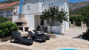 A white building with a stone chimney, a pool, and lounge chairs.