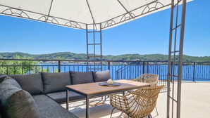 Un espacio al aire libre techado con sofá, mesa y sillas, con vista a un cuerpo de agua y colinas.