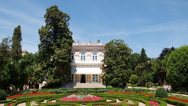 Ein formal angelegter Garten mit symmetrischer Anordnung, der von geometrisch geschnittenen Hecken und Blumenbeeten gesäumt wird und zu einem klassizistischen Gebäude mit Rundbogenfenstern und einem zentralen Eingang führt.