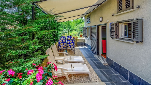 A covered outdoor seating area with chairs and a table, a building with shutters, and a garden with trees and flowers. - Karolina by Interhome (Lovran)