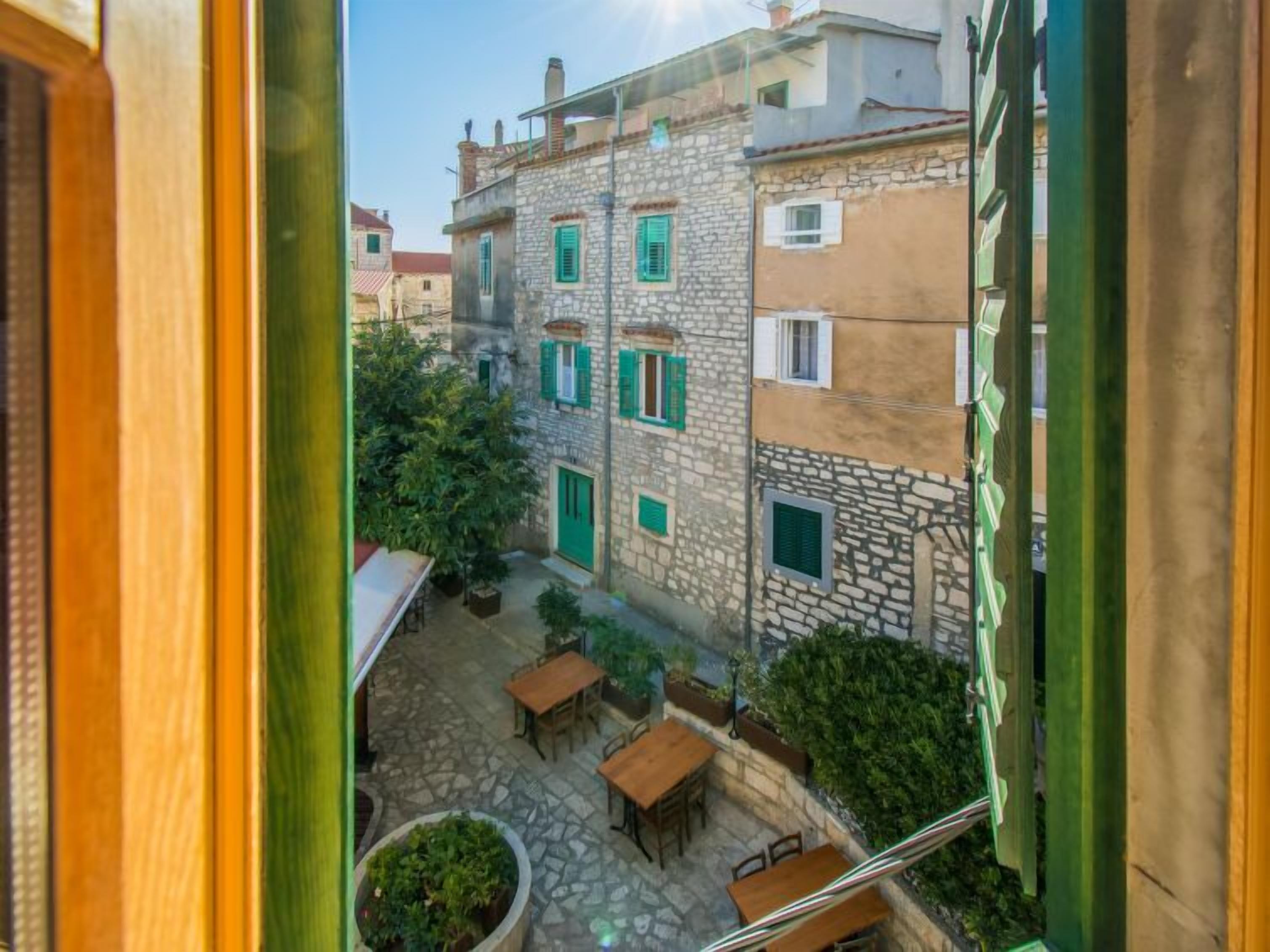 A courtyard with stone flooring, wooden tables, and green shutters on buildings.
