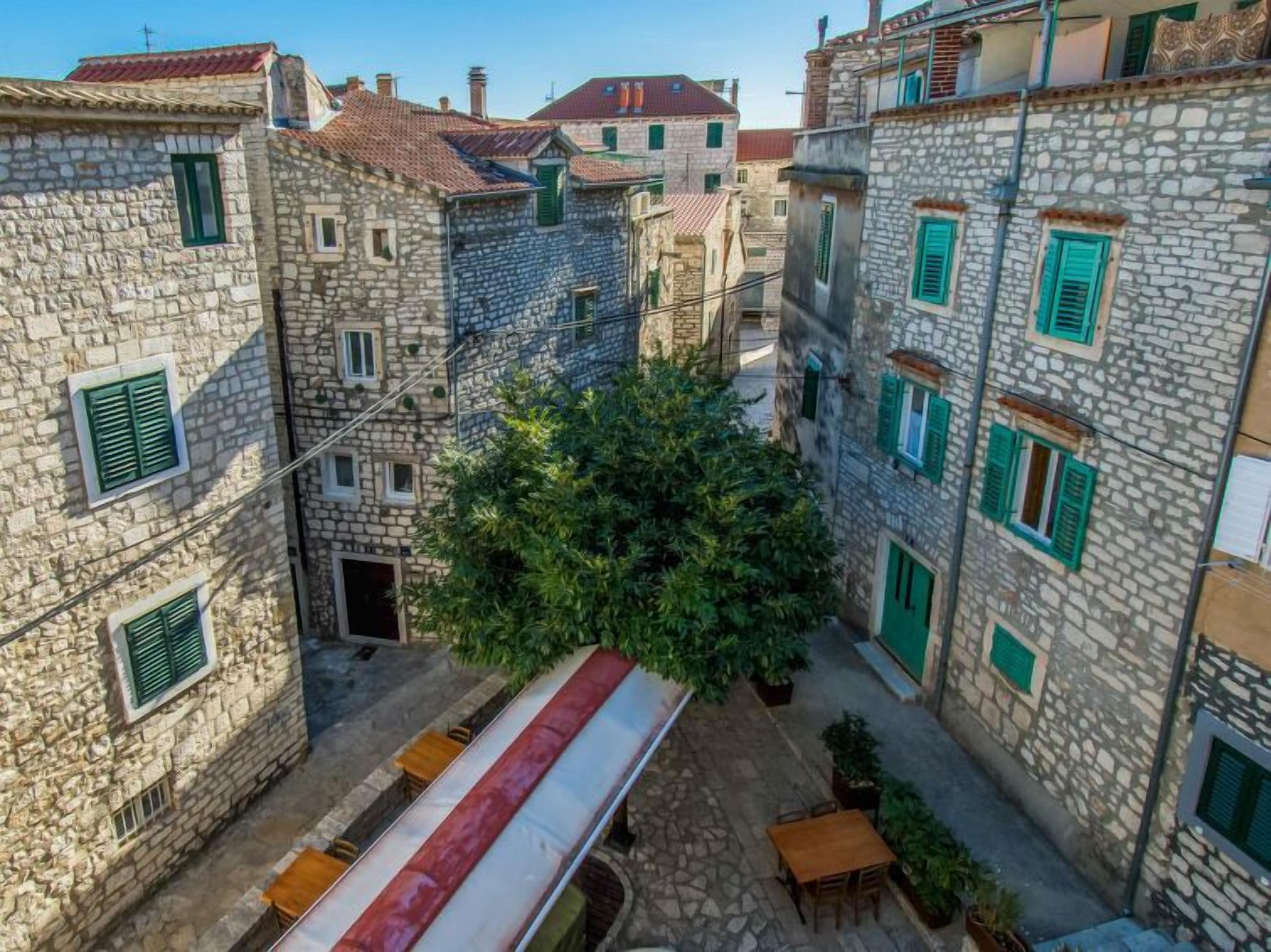 A narrow street with stone buildings, green shutters, and outdoor seating.