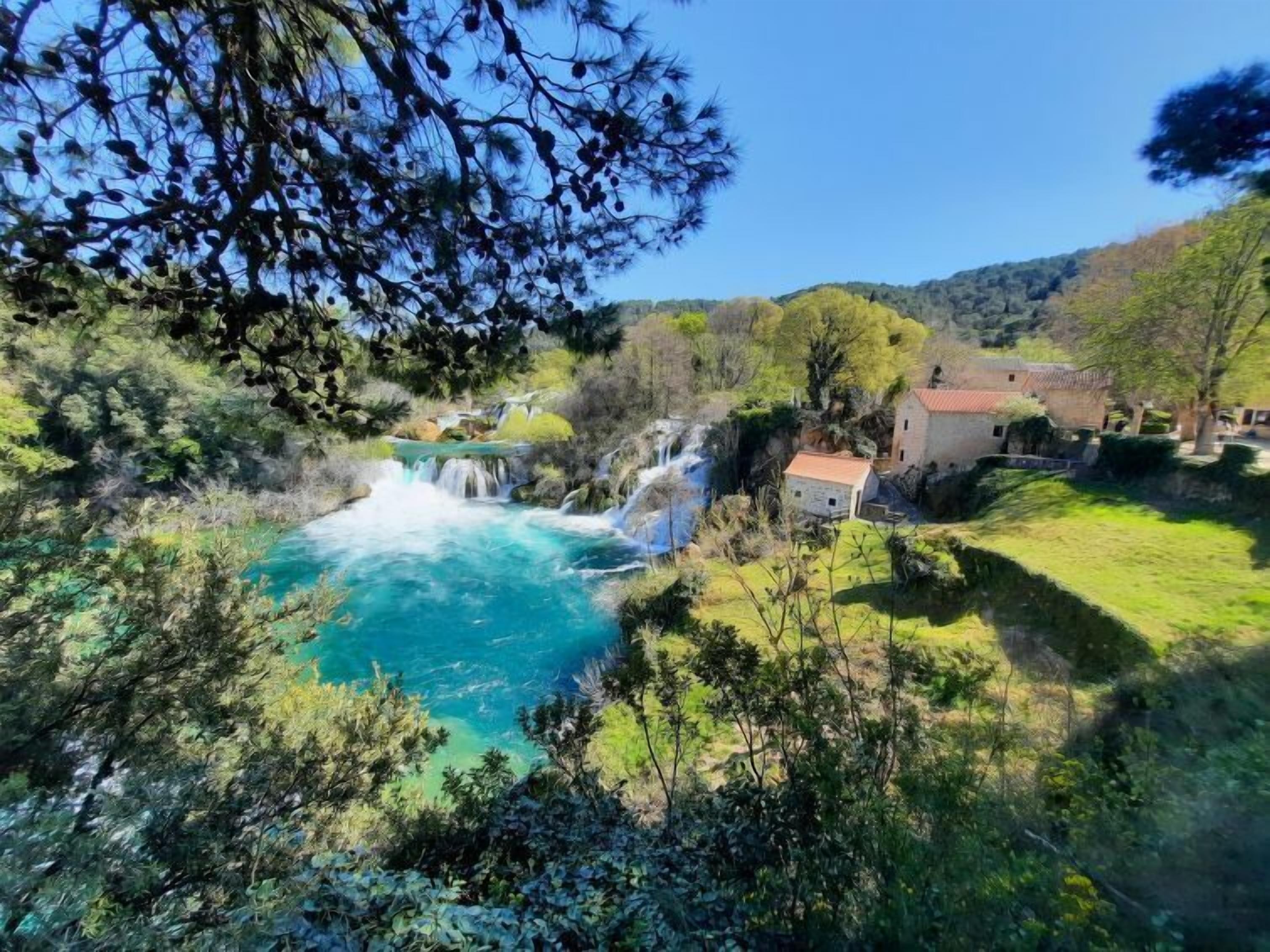 Eine natürliche Landschaft mit einem Wasserfall, einem Fluss und einem Gebäude im Hintergrund.