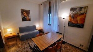 Une petite pièce chaleureuse, avec une table à manger en bois, deux chaises, un canapé, une petite table d’appoint et un lampadaire. Les murs sont ornés de posters et il y a une fenêtre avec des rideaux.