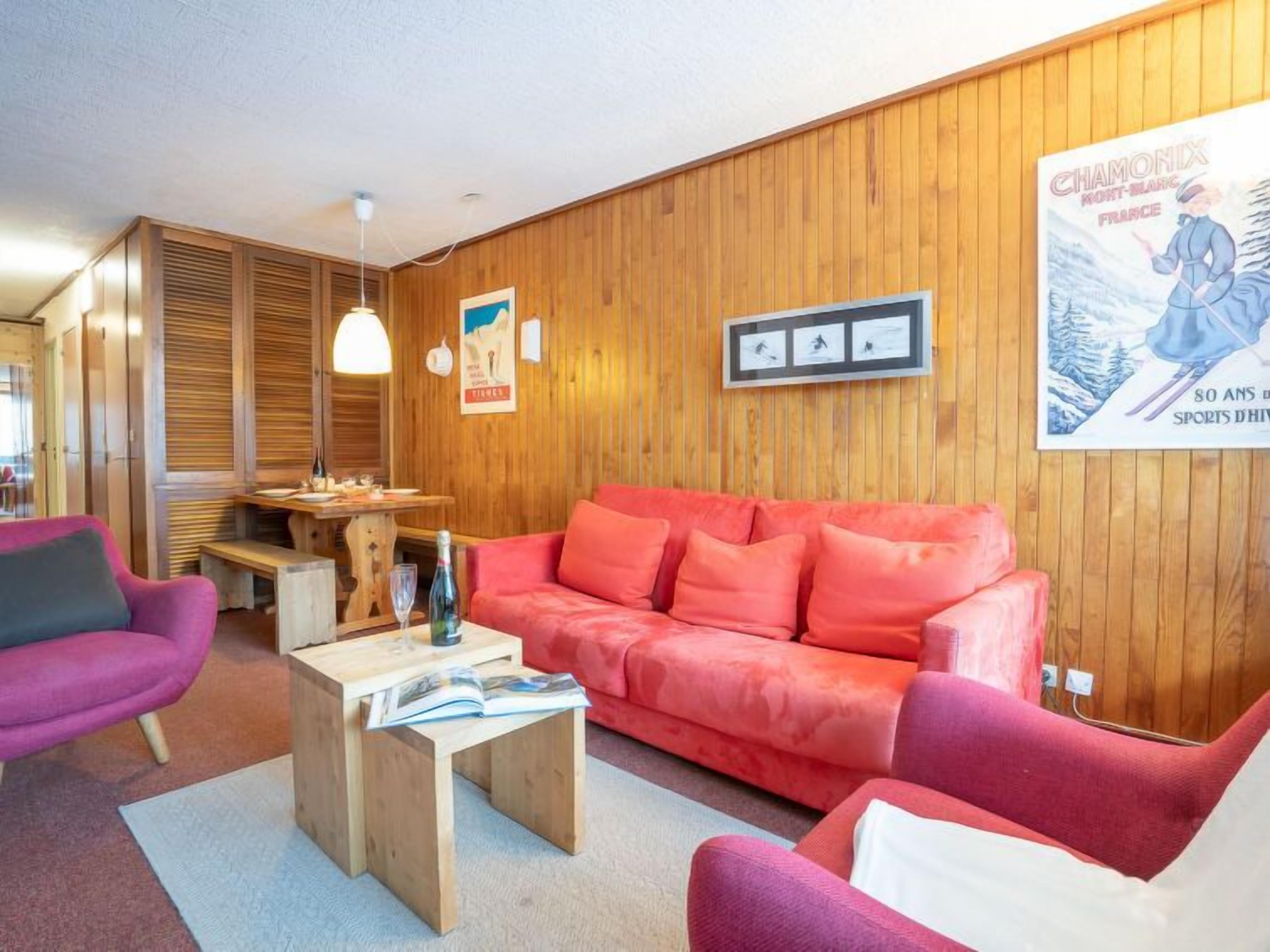 A cozy living room with a wooden wall, a red sofa, a pink armchair, and a wooden coffee table with a magazine.