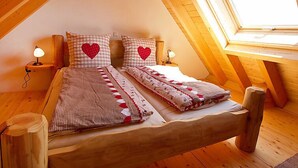 A cozy attic bedroom with a sloped ceiling, wooden beams, a large window, and a bed with checkered bedding and heart-shaped pillows.