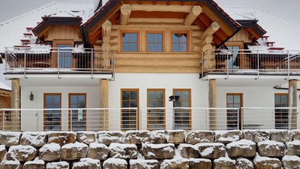 A two-story log cabin with a snow-covered roof and stone wall foundation.