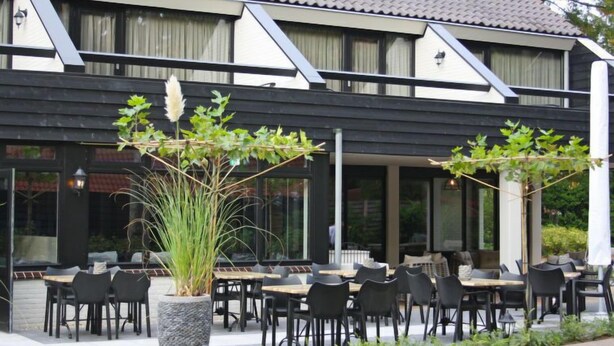 An outdoor seating area with tables and chairs, a large planter with plants, and a building with a tiled roof and windows. - Vacation home 2+ by Interhome (Lochem)