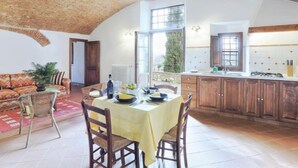 Une salle à manger spacieuse avec un haut plafond et des poutres en bois rustiques, un coin salon confortable avec un canapé à motifs, et une cuisine dotée de meubles en bois et d’une grande fenêtre.