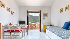 Una habitación con una cama, un escritorio, una silla, una mesa con un cojín rojo y vistas a una montaña a través de una puerta abierta.