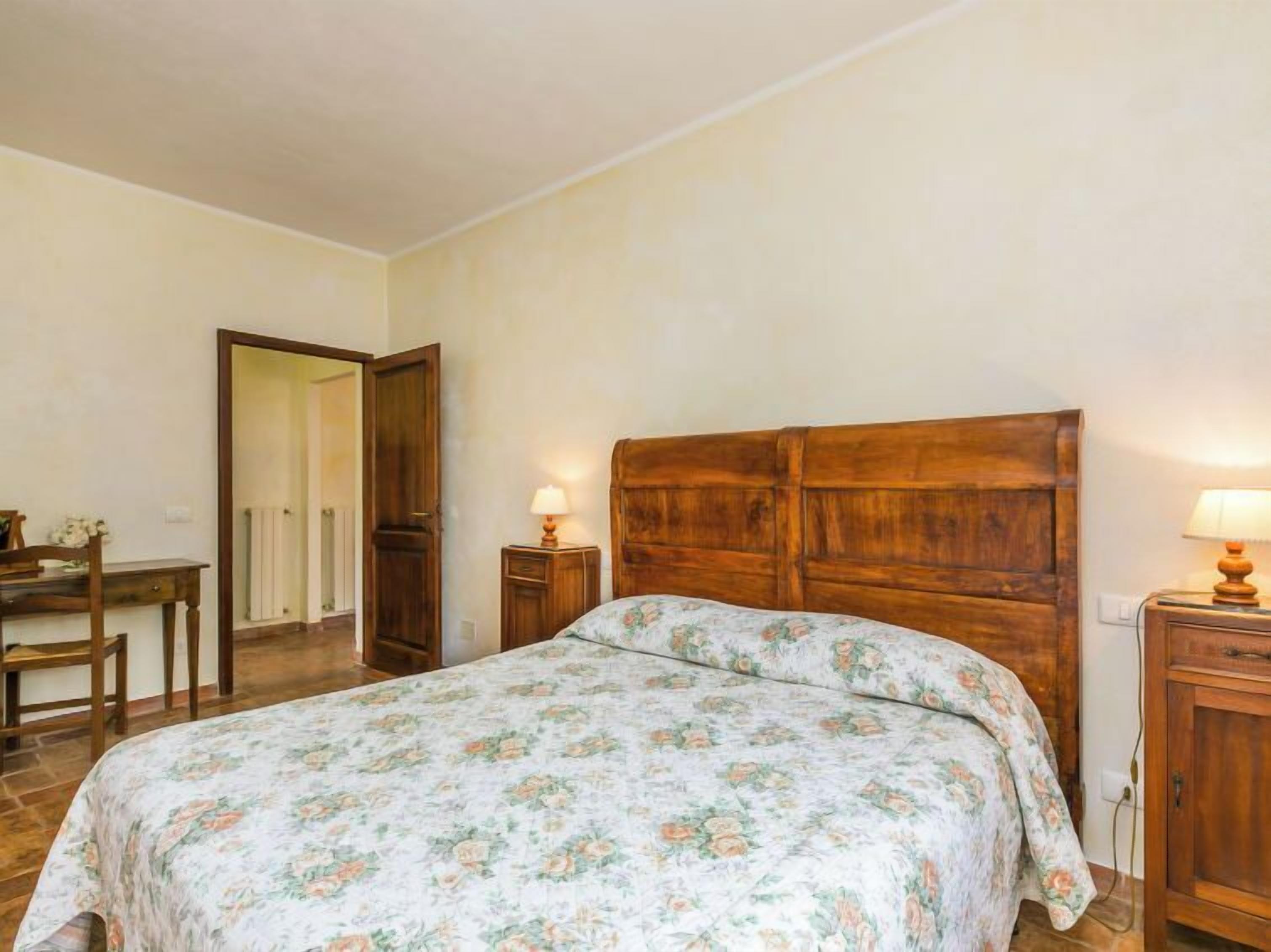A bedroom with a wooden headboard, a floral bedspread, a bedside table with a lamp, and a wooden nightstand.