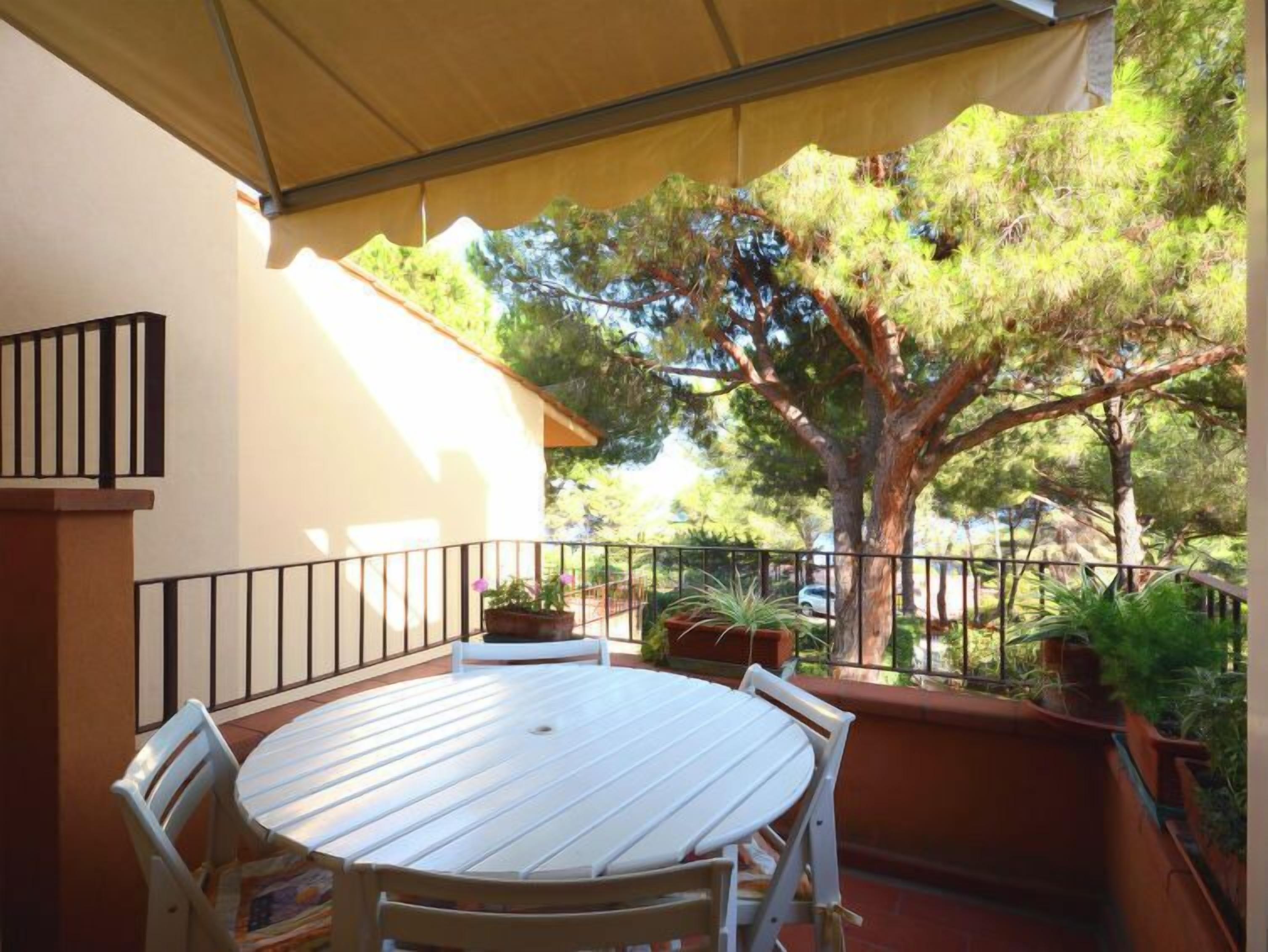 Un patio cubierto con una mesa redonda y sillas, rodeado de plantas en macetas y un árbol grande.
