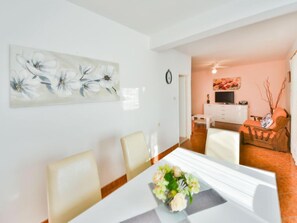 Un coin repas avec une table blanche, des chaises et un vase de fleurs. Il y a un tableau au mur et une horloge.