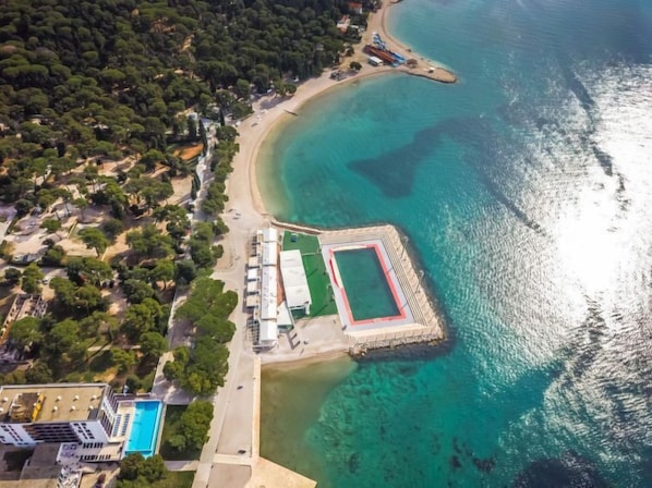 Vue aérienne d’une zone côtière comprenant une plage, une piscine et un bâtiment à la structure longue et étroite.