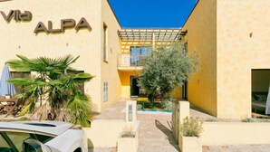 Ein Gebäude mit der Beschilderung 'Vila ALPA', das einen zentralen Innenhof mit einer Pergola, Topfpflanzen und einem geparkten Auto vor der Fassade zeigt.