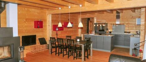 A spacious living area with a fireplace, wooden flooring, a dining table with chairs, a sofa, and a kitchen in the background.