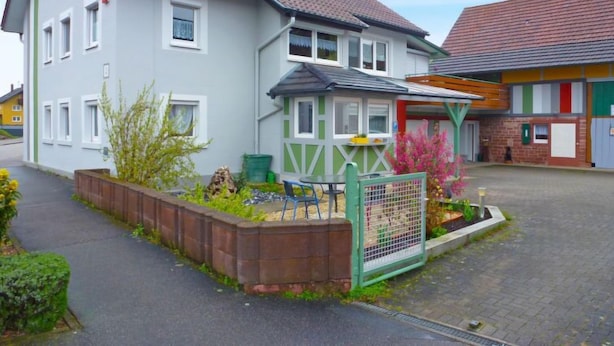 A two-story house with a grey facade, a tiled roof, and a small garden area with plants and a bench. - Apartment Bless by Interhome (Schutterzell)