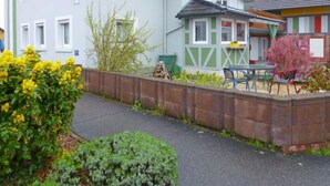 A two-story building with a grey facade, multiple windows, and a small garden area with plants and a table with chairs. - Apartment Bless by Interhome (Schutterzell)