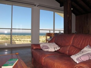 Un sofá de cuero con cojines estampados, una mesa de centro de madera con un libro, y vista a la playa a través de amplias ventanas.