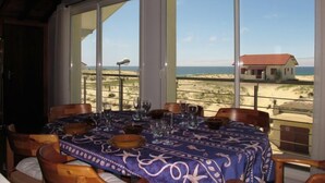 A dining area with a table set for four, large windows offering a view of the beach and ocean, and a hanging light fixture above. - Apartment Capama by Interhome (Mimizan)