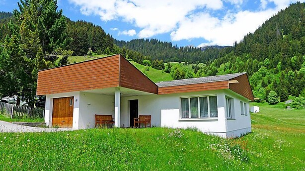 Une maison moderne à deux étages, dotée d’un toit en pente, d’une porte d’entrée en bois et d’un petit porche avec deux chaises.