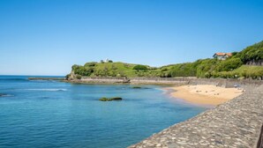 Escena costera con un sendero de piedra que lleva a una playa de arena, una colina cubierta de césped con un edificio y un cielo azul despejado.