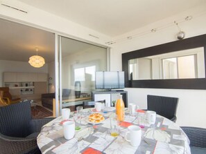 Un coin repas avec une table dressée pour deux personnes, un téléviseur en arrière-plan et une porte-fenêtre coulissante donnant sur la cuisine.