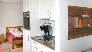 Een compacte slaapkamer met een bed, nachtkastje en een kleine bedlamp. Grenzend aan de slaapkamer bevindt zich een kitchenette met een fornuis, koffiezetapparaat en een houten plank.