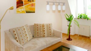 Een woonkamer met een bank, een bijzettafeltje, een vloerlamp, een schilderij, een raam met planten en een radiator.