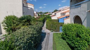 Un étroit sentier serpente entre des bâtiments aux murs blancs et aux toits recouverts de tuiles rouges, le tout étant bordé de verdure.