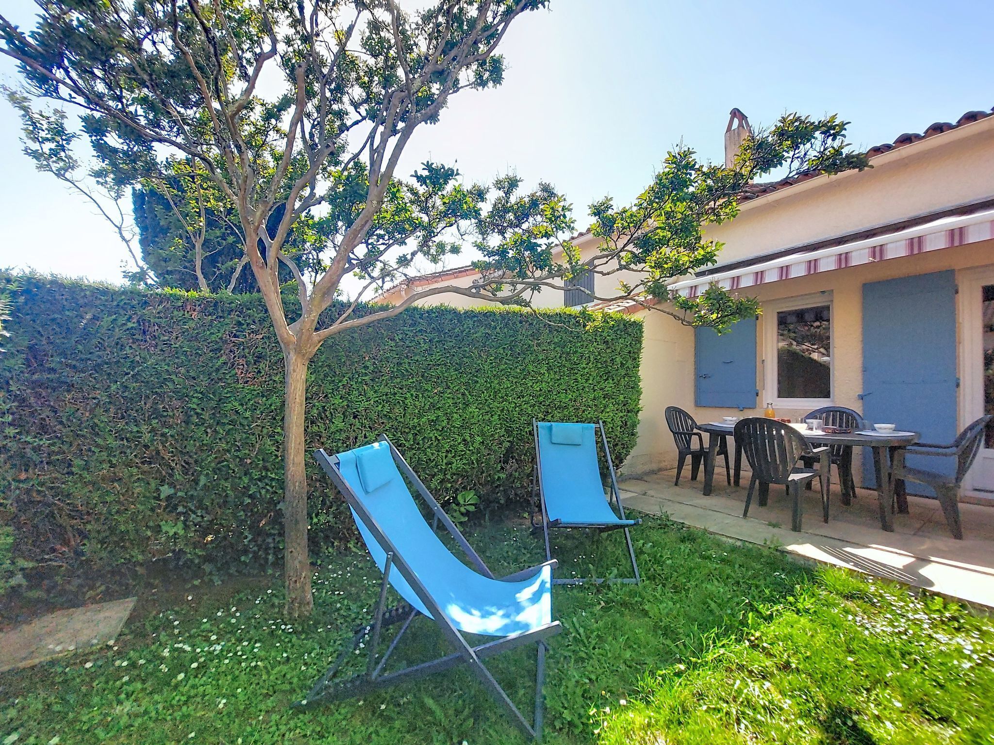 Une terrasse avec deux chaises bleues, une table et un petit espace jardin agrémenté d’un arbre et d’une haie.