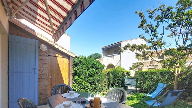 Un espace de restauration en plein air avec une table dressée pour deux personnes, entourée de chaises, et une vue sur un bâtiment et de la verdure en arrière-plan.