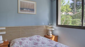 A bedroom with a bed, a nightstand, a window with a view of greenery, and a framed picture on the wall.