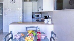 A modern kitchen with white cabinets, a microwave, and a coffee maker. There is a dining table with chairs and a variety of fruits and beverages.