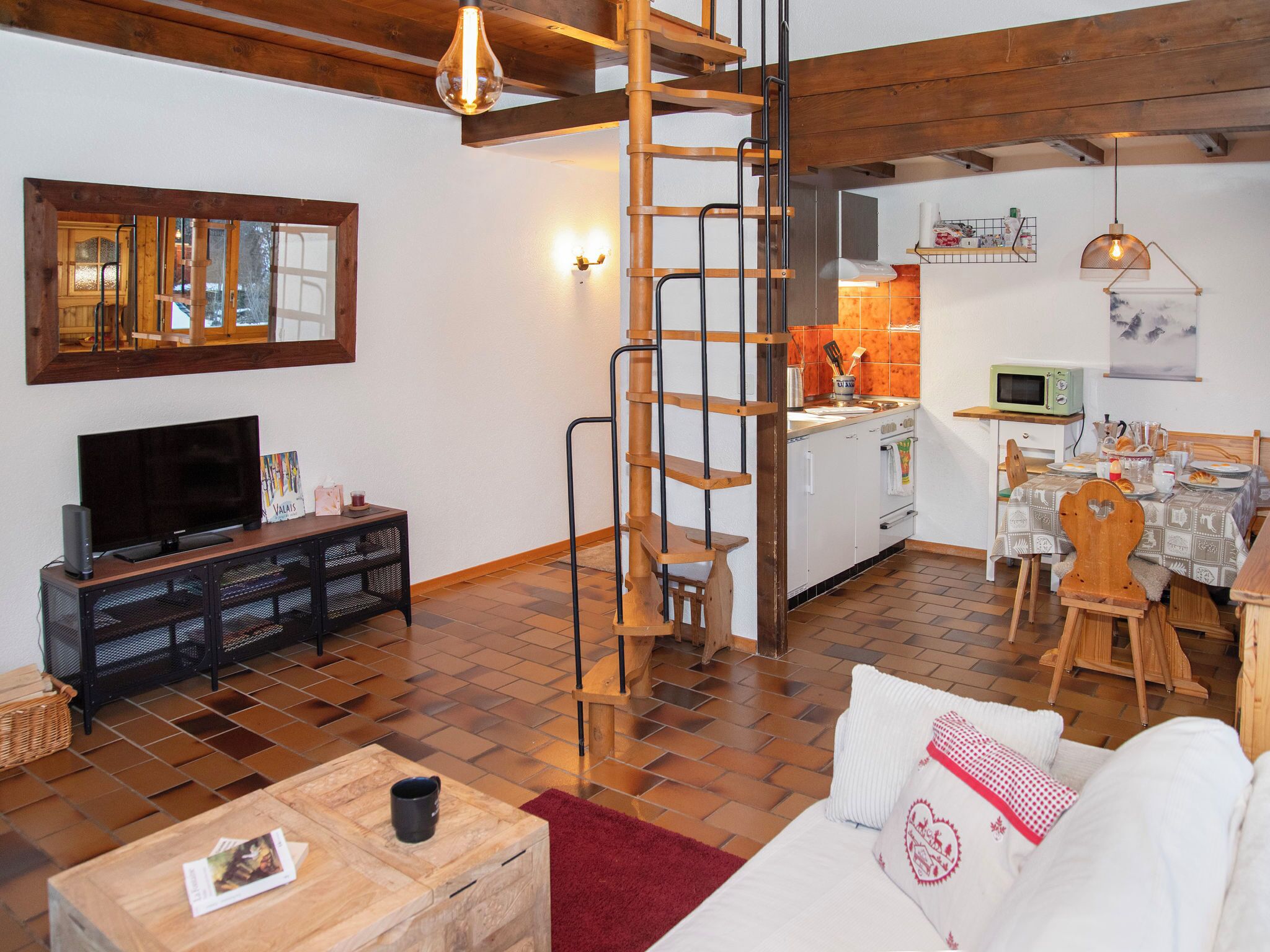 Un salon chaleureux avec un escalier en bois, un téléviseur et un coin repas, avec une cuisine en arrière-plan.