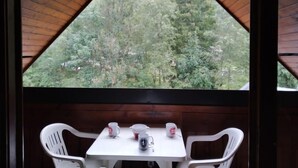 A wooden cabin with a triangular roof and a table with chairs. A window reveals a forested mountain view. - Pierres Blanches F et H by Interhome (Les Contamines)