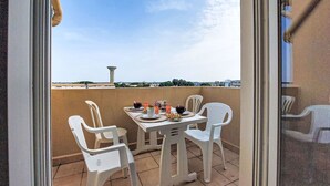 Un balcon avec une table dressée pour deux personnes, des chaises en plastique blanches et une vue sur le paysage urbain et une tour au loin.