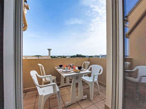 Un balcon avec une table dressée pour deux personnes, des chaises en plastique blanches et une vue sur le paysage urbain et une tour au loin.