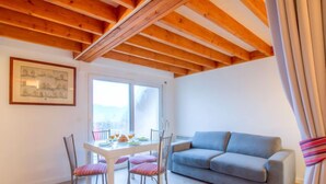 A cozy room with a wooden ceiling, a dining table with chairs, a grey sofa, and a large window offering a view. - Les Reflets de Carnac by Interhome (Carnac)