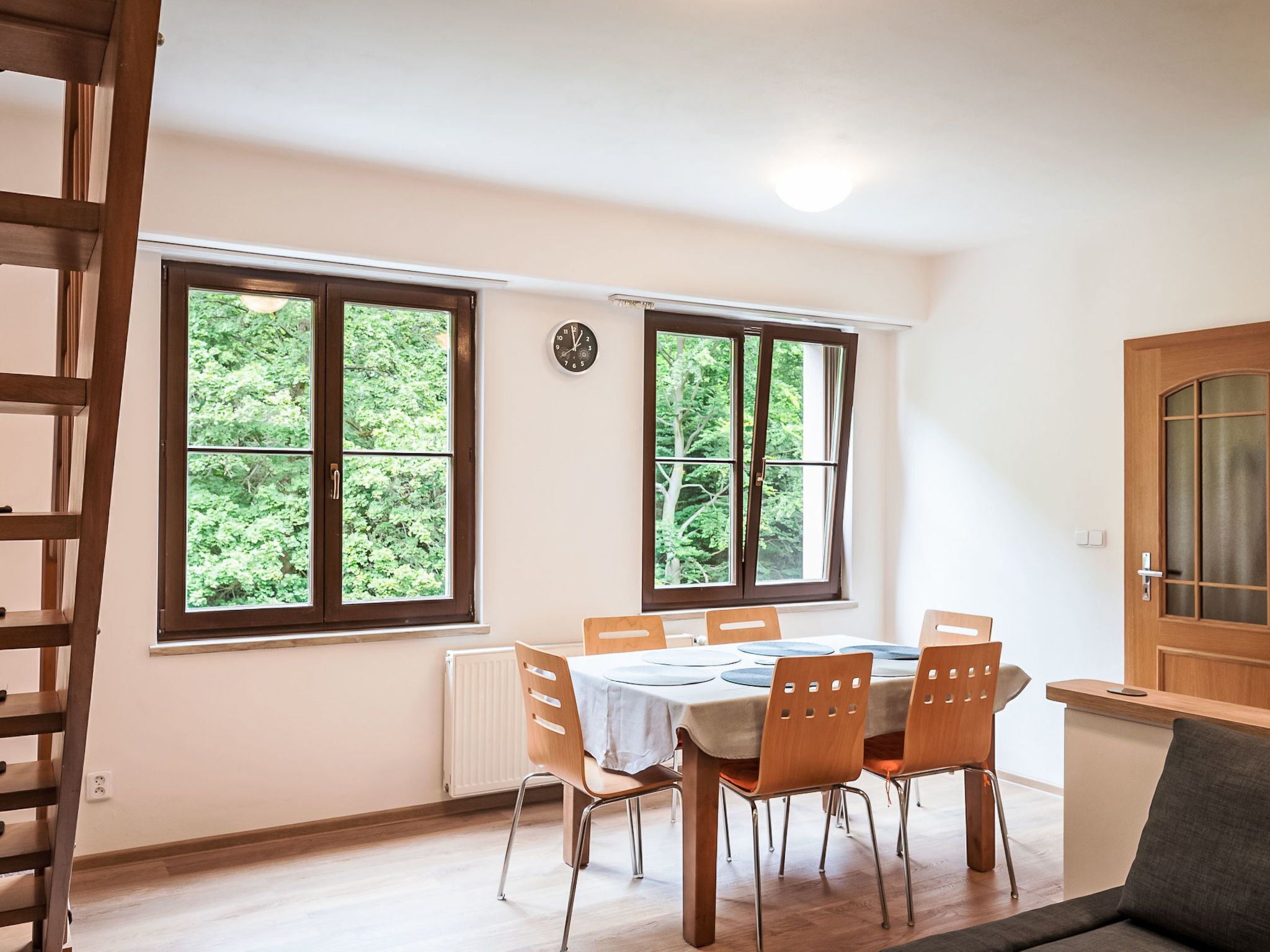 Un coin repas avec une table dressée pour quatre personnes, deux fenêtres donnant sur un paysage verdoyant, une horloge au mur et une porte en bois.
