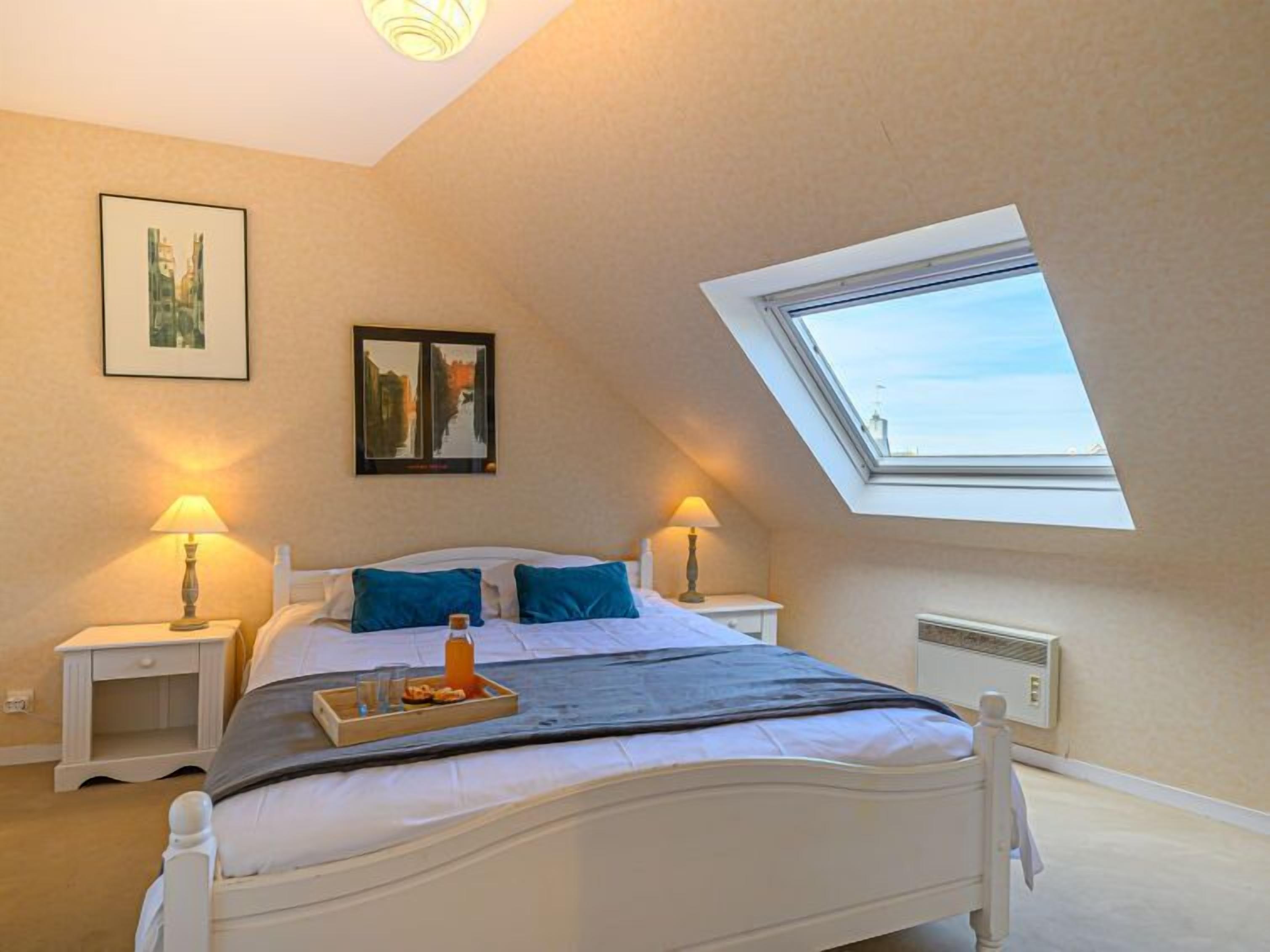 A bedroom with a skylight, a bed with pillows, bedside tables with lamps, and artwork on the walls.