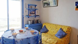 Una habitación con una mesa de comedor lista para dos comensales, un sofá amarillo con almohadas azules, una pared azul con estantes que exhiben diversos objetos y un cuadro en la pared.