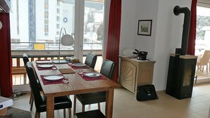 A dining area with a wooden table set for four, a kitchenette with a stove, and a view of buildings through large windows. - Promenade Wohnung 21 by Interhome (Davos)